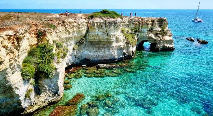 Scogliere bianche e mare cristallino sul litorale adriatico. Turisti ammirano il panorama, un catamarano naviga in lontananza.