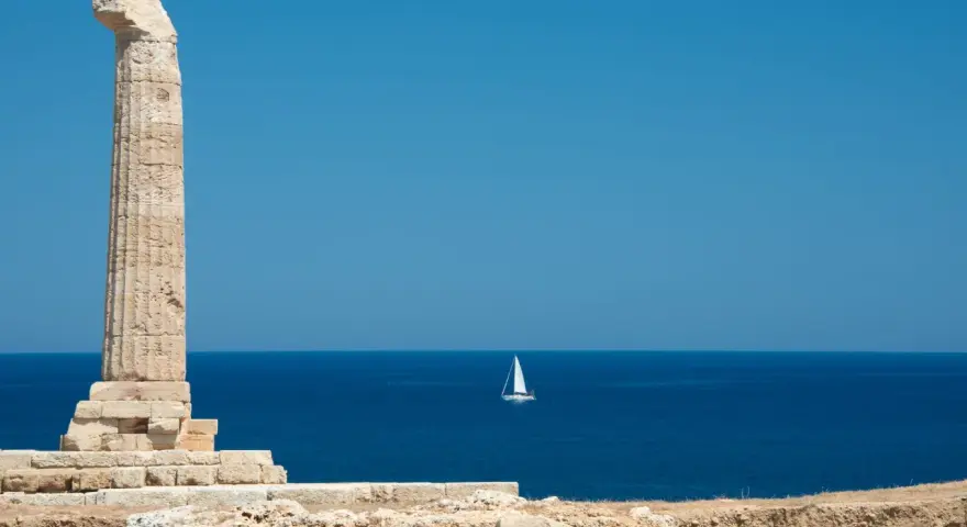 Antica colonna sulla costa degli Achei, con un veliero solitario all'orizzonte sotto un cielo azzurro.
