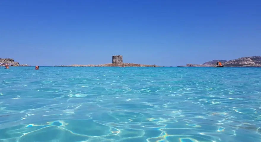Acque cristalline e un antico torrione su un'isola: un mare bello in Italia, con gente che nuota e una barca a vela all'orizzonte.