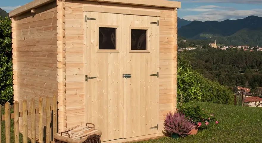 La casetta in campagna, in legno chiaro, con due finestrelle e un cesto di legna, si affaccia su un panorama collinare con un borgo in lontananza.