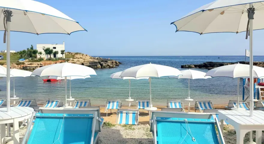 Lido Cala Pescatore: lettini azzurri e ombrelloni bianchi su una spiaggia di ciottoli, con il mare cristallino e rocce sullo sfondo.