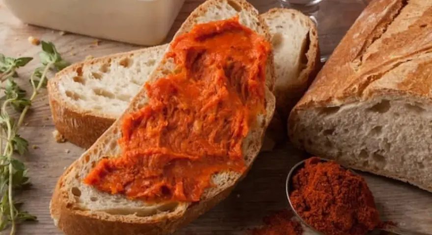 Fetta di pane con 'nduja, uno dei piatti tipici della Calabria, accanto a una ciotola con la crema e un cucchiaino di peperoncino in polvere.