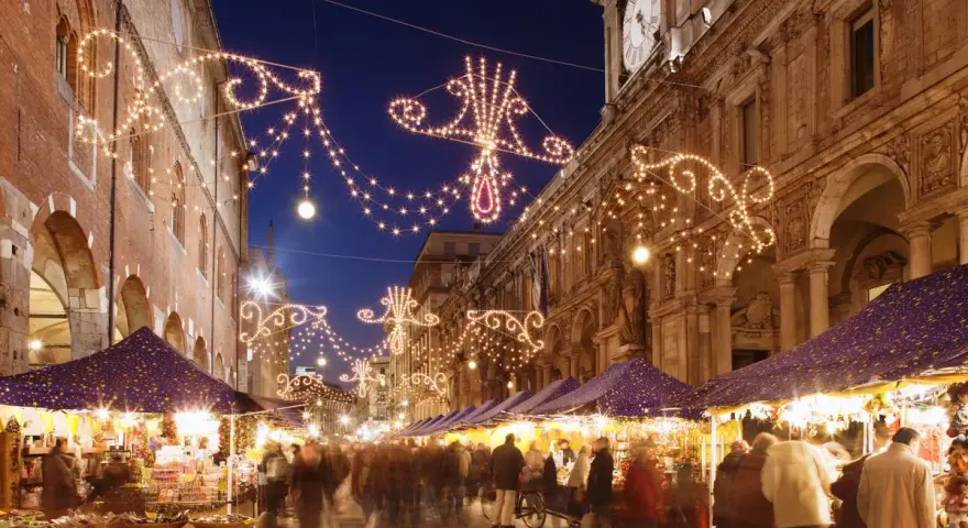 Una strada illuminata da lucine natalizie, con bancarelle e gente che passeggia: ecco i mercatini di Natale più belli.