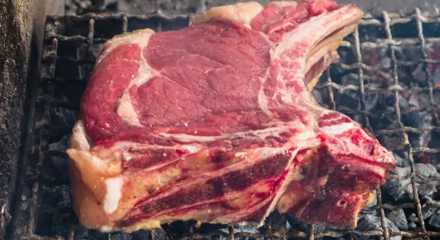 Una succulenta fiorentina di vitello, con la sua succosa carne rossa e il grasso invitante, cuoce lentamente sulla griglia ardente, sprigionando un profumo delizioso.