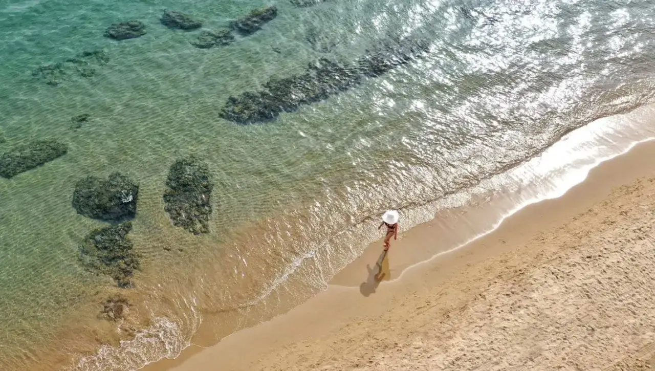 Una donna con cappello bianco cammina su una delle splendide spiagge del Lazio, tra sabbia dorata e acque cristalline con rocce sommerse.