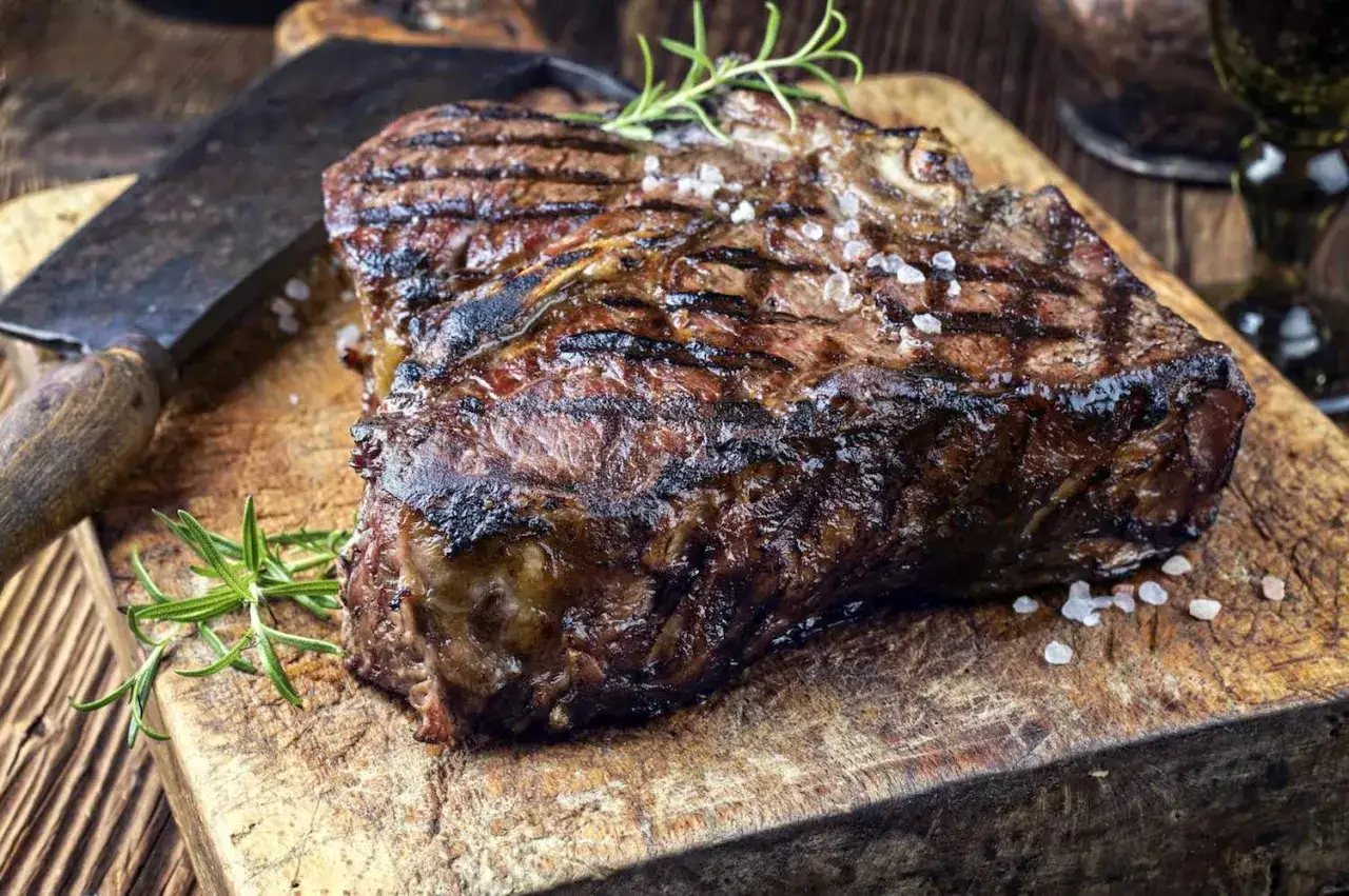 Una succulenta fiorentina di vitello, grigliata alla perfezione, adagiata su un tagliere rustico con rosmarino e sale grosso.