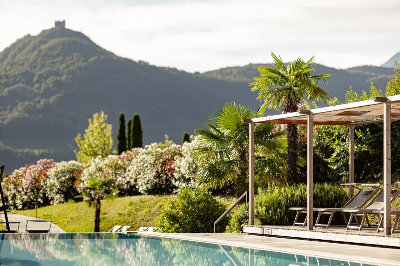 Piscina e lettini sotto un pergolato al hotel Weingarten Kaltern, con vista su colline fiorite e un castello in lontananza.