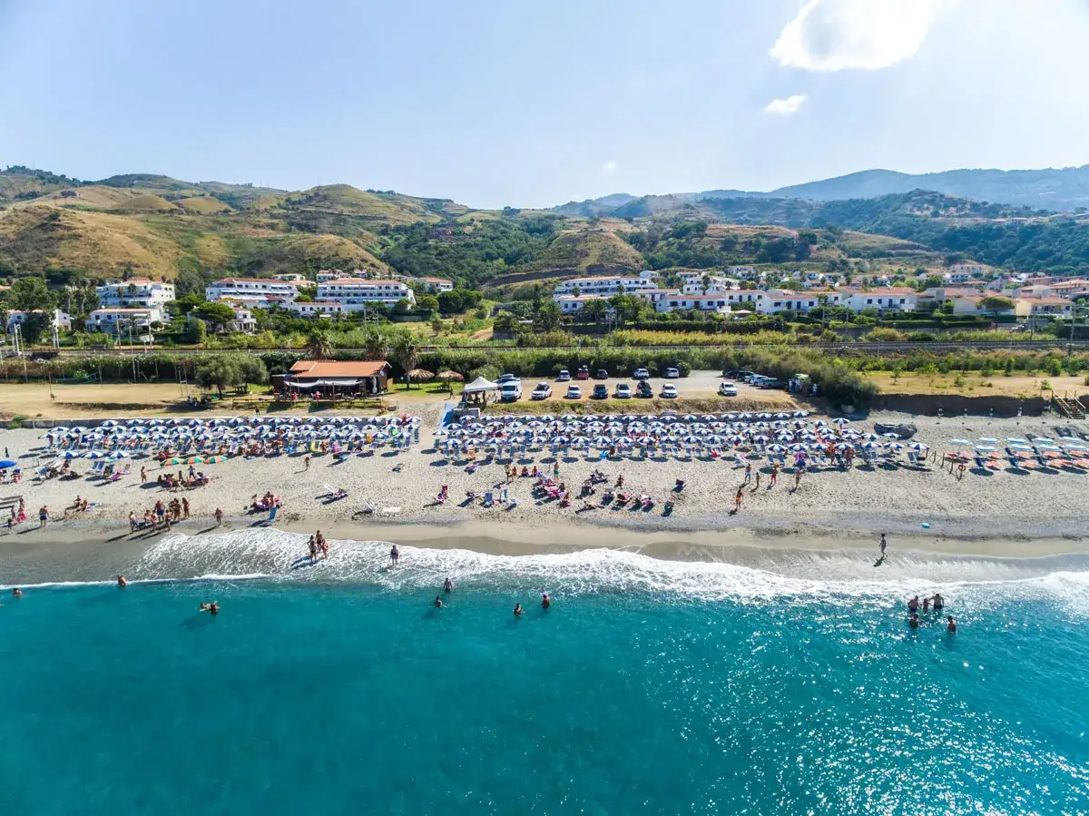 Spiaggia affollata con ombrelloni e lettini, mare azzurro e colline verdi sullo sfondo. Il hotel club residence Martinica offre una vacanza da sogno.