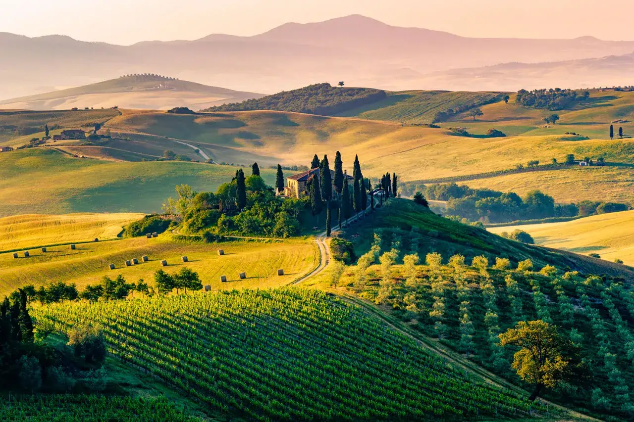 Dolci colline toscane con vigneti, cipressi e un casale in cima, sotto un cielo rosato. Un paesaggio da cartolina delle città toscane.