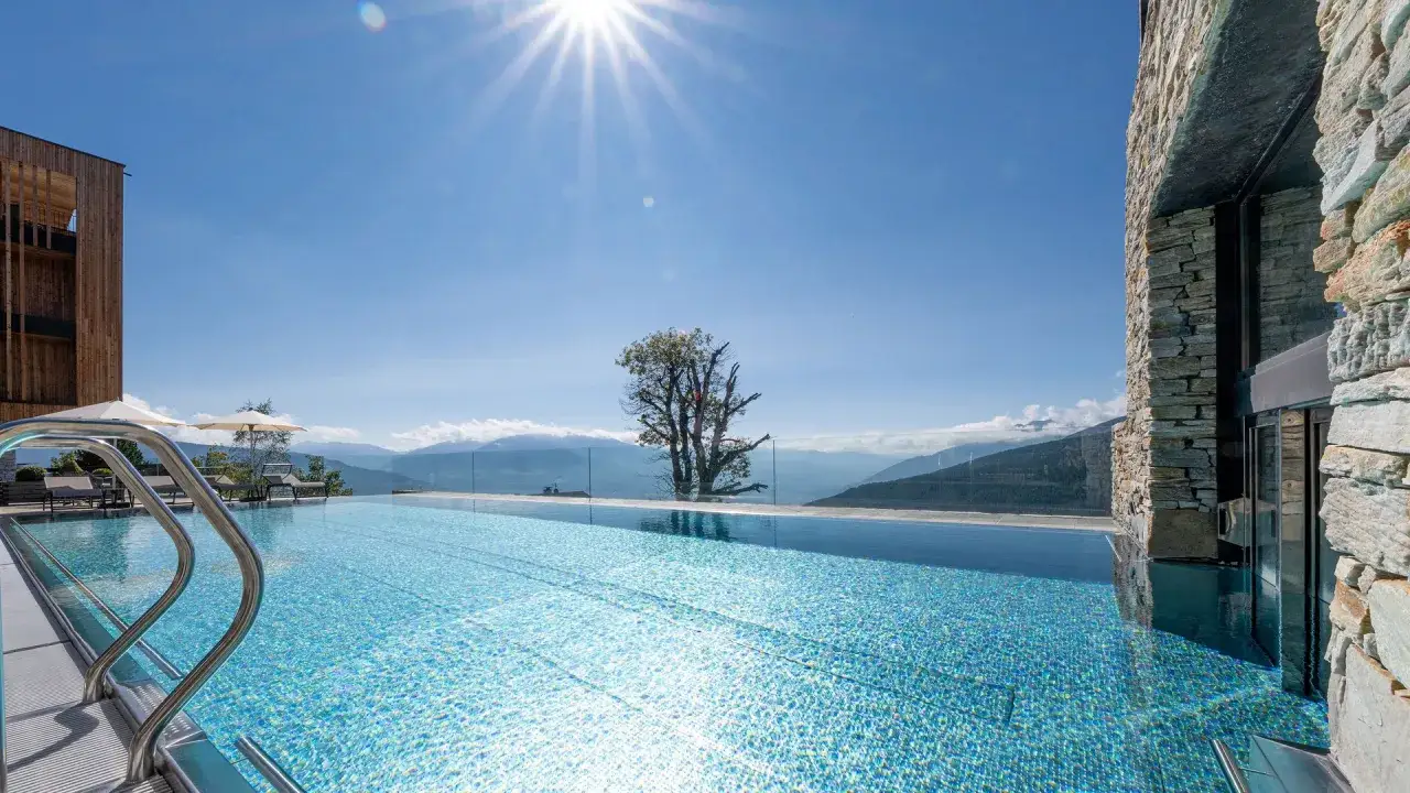 Piscina a sfioro con vista panoramica sulle montagne, parte dell'hotel Milla Montis. Sole splendente, cielo azzurro, architettura moderna e naturale.