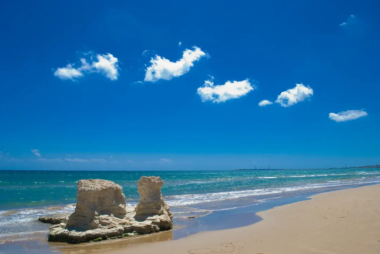 Scogliere calcaree e sabbia dorata sulle spiagge Sicilia orientale, sotto un cielo azzurro con nuvole bianche.