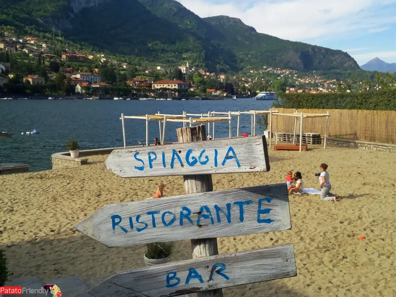 Segnaletica per spiaggia, ristorante e bar su una sabbia dorata, con montagne verdi e laghi balneabili sullo sfondo.