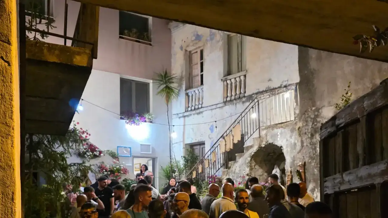 Serata estiva a Oliena, Sardegna. Un cortile illuminato da lucine, con persone che chiacchierano tra fiori e architetture tipiche.