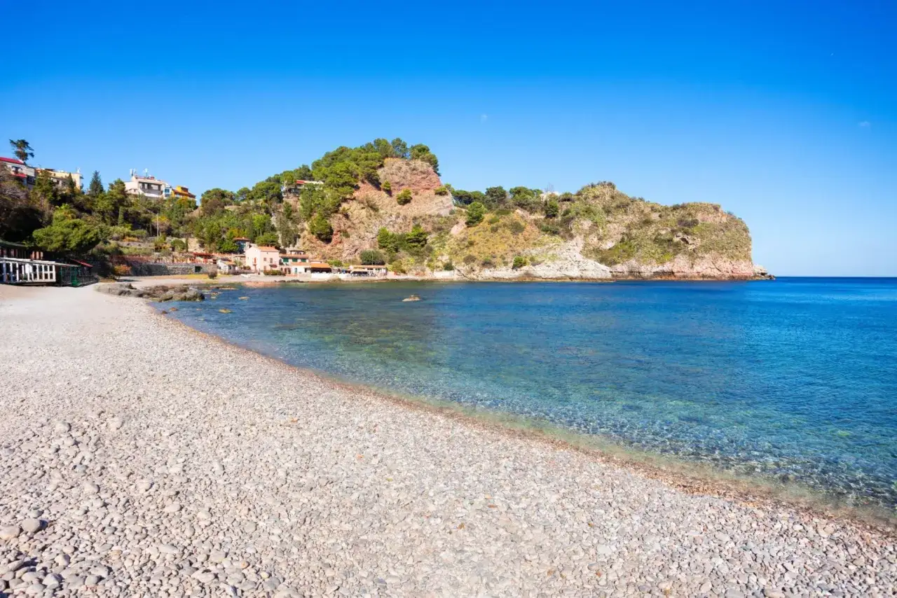 Spiaggia di ciottoli con acque cristalline e case arroccate sulla collina. Un luogo incantevole tra i tanti posti da visitare in Sicilia.