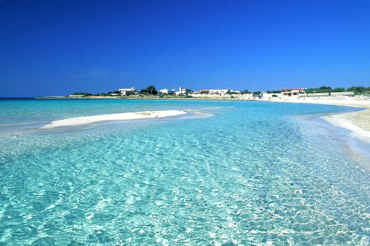Acque cristalline e sabbia bianca a Punta Prosciutto, le Maldive del Salento, con case bianche sullo sfondo.