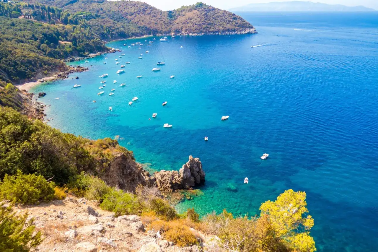 Baia con acque cristalline e numerose barche, tipica del Grosseto mare. Vegetazione lussureggiante e scogliere a picco sul mare.