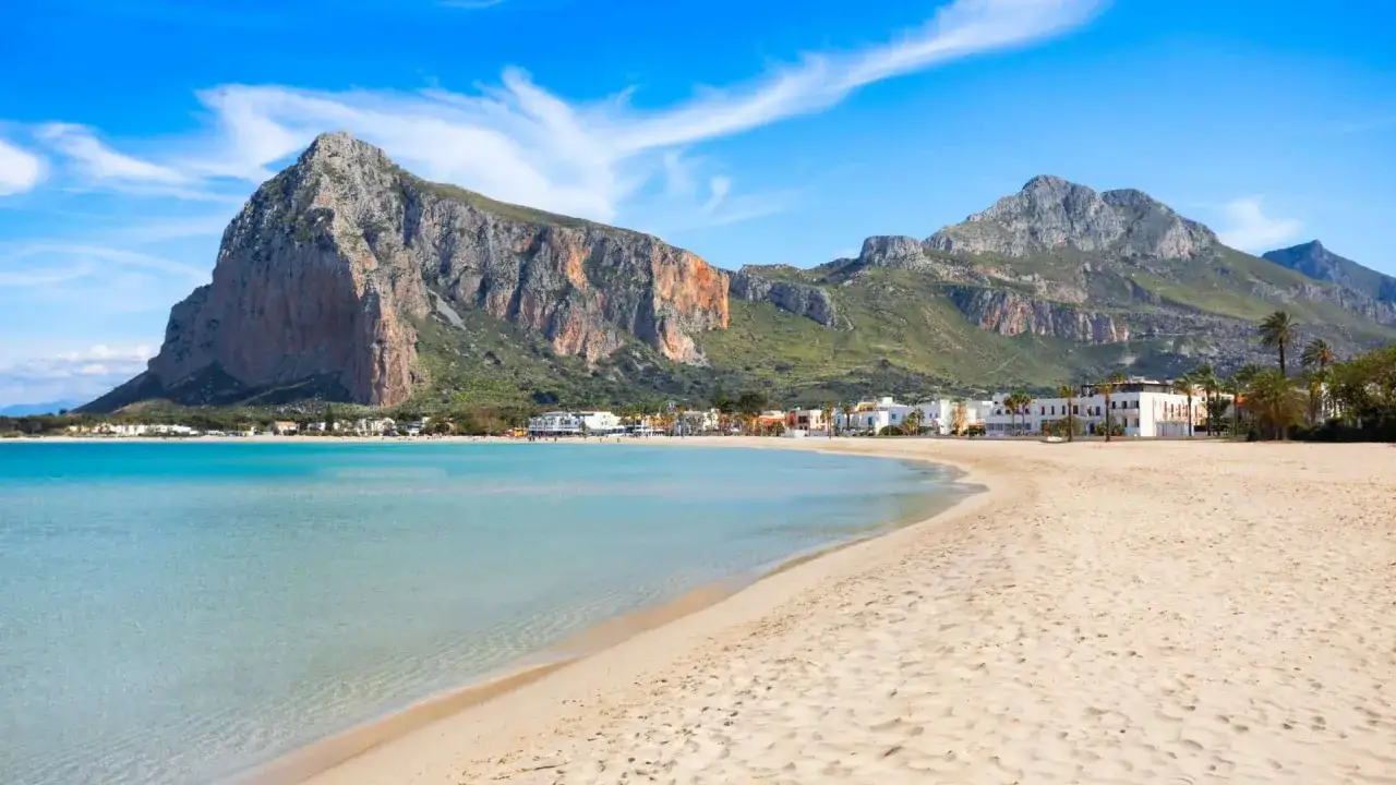 Spiaggia dorata e mare cristallino in Sicilia, con imponenti montagne sullo sfondo e un villaggio costiero.