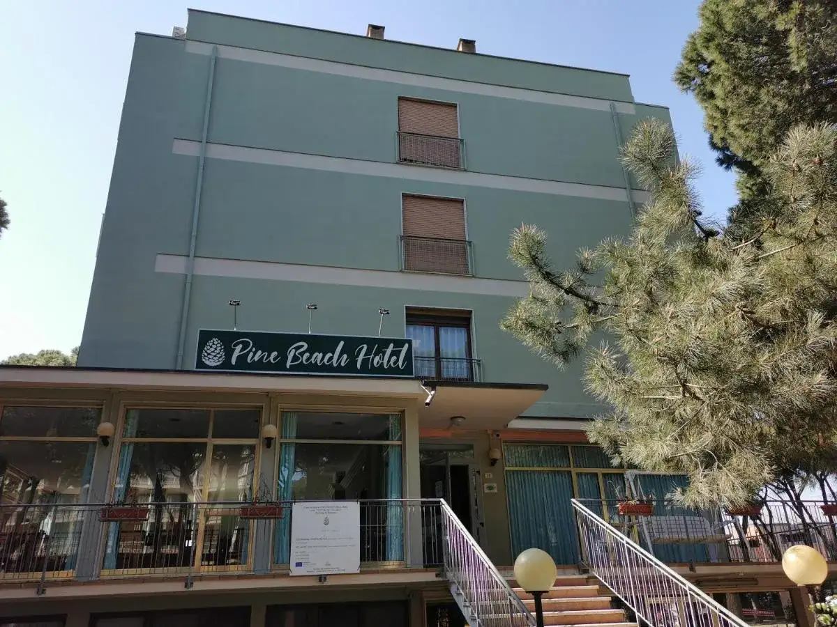 Il Pine Beach Hotel a Rosolina Mare, con la sua facciata verde acqua e le finestre ampie, è circondato da pini rigogliosi.