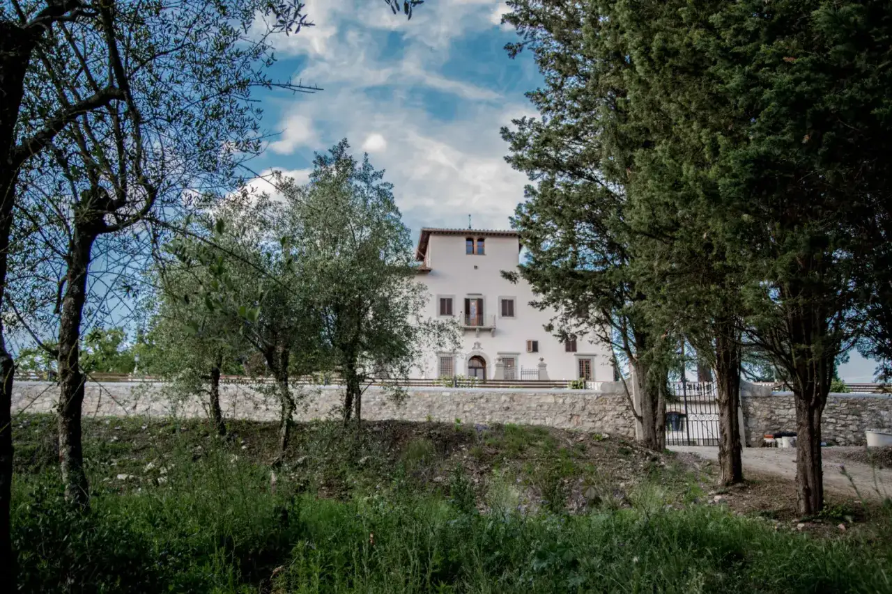 La villa d'arte Pontassieve, circondata da alberi e vegetazione, si staglia contro un cielo azzurro con nuvole bianche.
