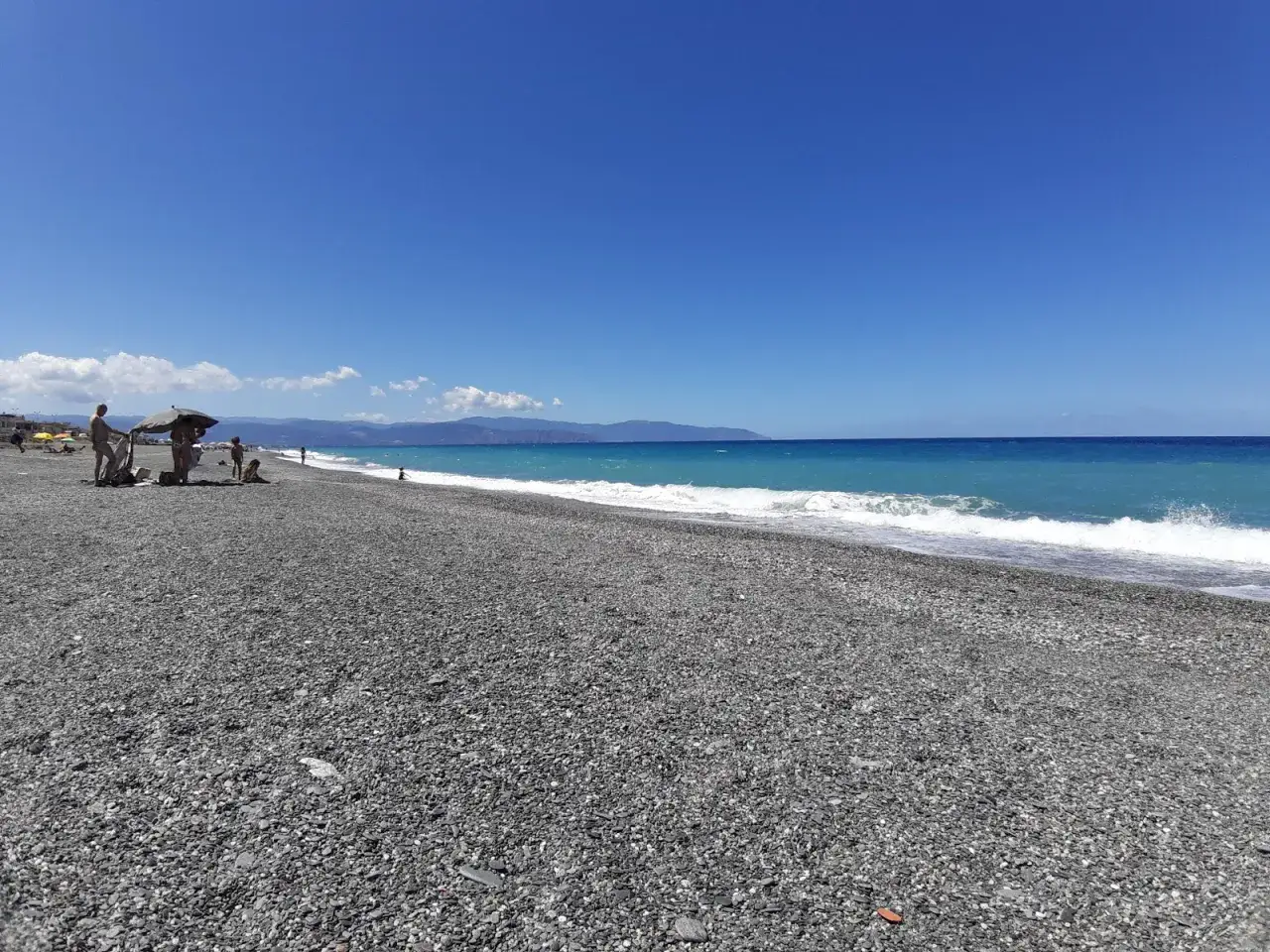 Spiaggia delle Gole dell'Alcantara con ciottoli scuri, mare azzurro e montagne sullo sfondo. Famiglie si godono il sole.
