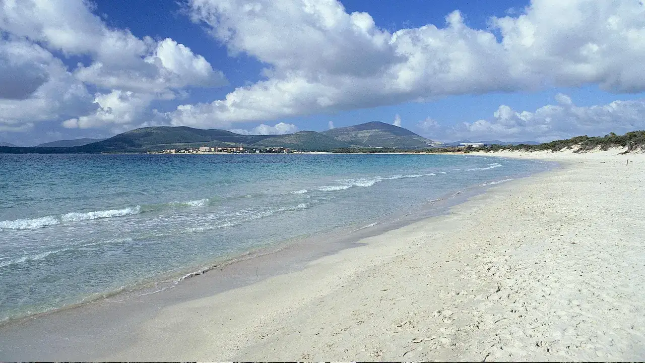 Spiaggia di Fertilia con mare cristallino, sabbia bianca e colline verdi all'orizzonte sotto un cielo azzurro con nuvole bianche.