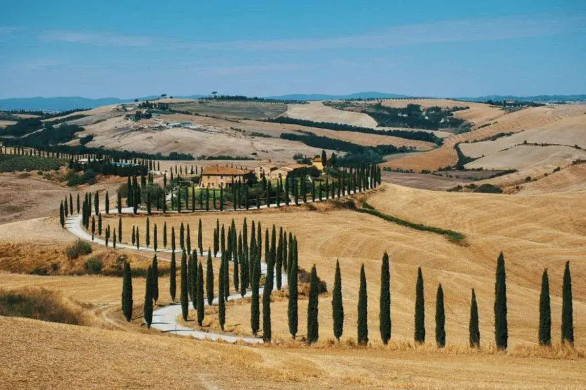 Un casale toscano circondato da cipressi lungo le strade del vino toscana. Campi dorati e colline ondulate sotto un cielo azzurro.