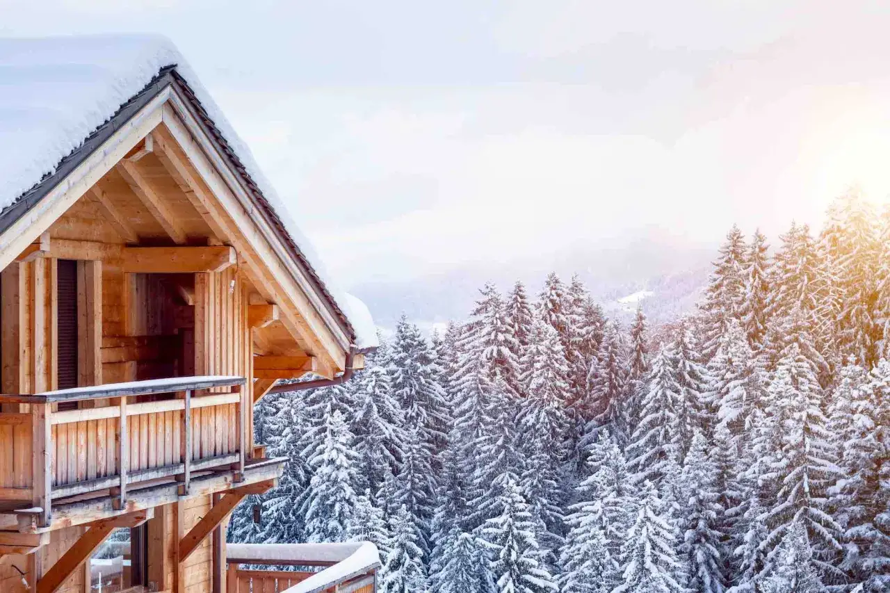 La piccola baita in legno, coperta di neve, si affaccia su un bosco di abeti innevati, con il sole che filtra tra i rami.