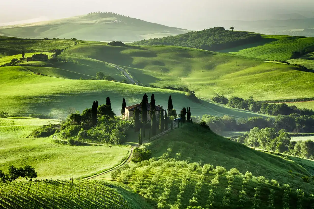 Colline verdi ondulate in Toscana, con una casa in pietra e cipressi su una collina. Un sogno per un viaggio in Toscana.