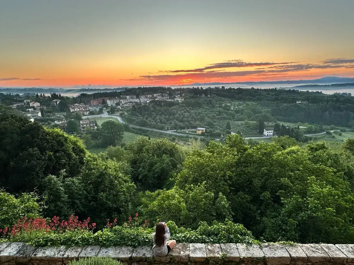 Alba in Toscana, con una persona che ammira il panorama dalla sua casetta in campagna. Colline verdi, un borgo e un cielo infuocato.