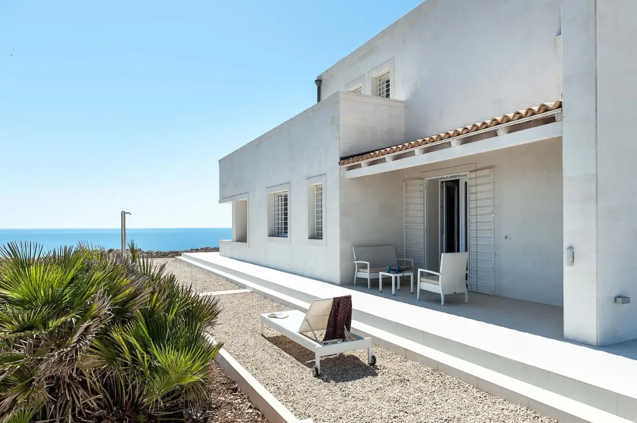 Pensione Scala Portopalo: moderna villa bianca con vista mare, lettino prendisole e vegetazione mediterranea.
