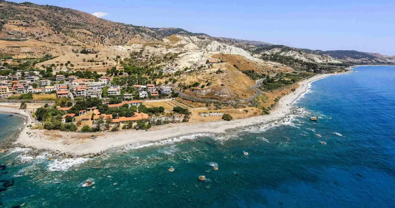 Paesaggio mozzafiato sulla costa degli Achei: case con tetti rossi, spiaggia sassosa e mare blu intenso sotto un cielo sereno.