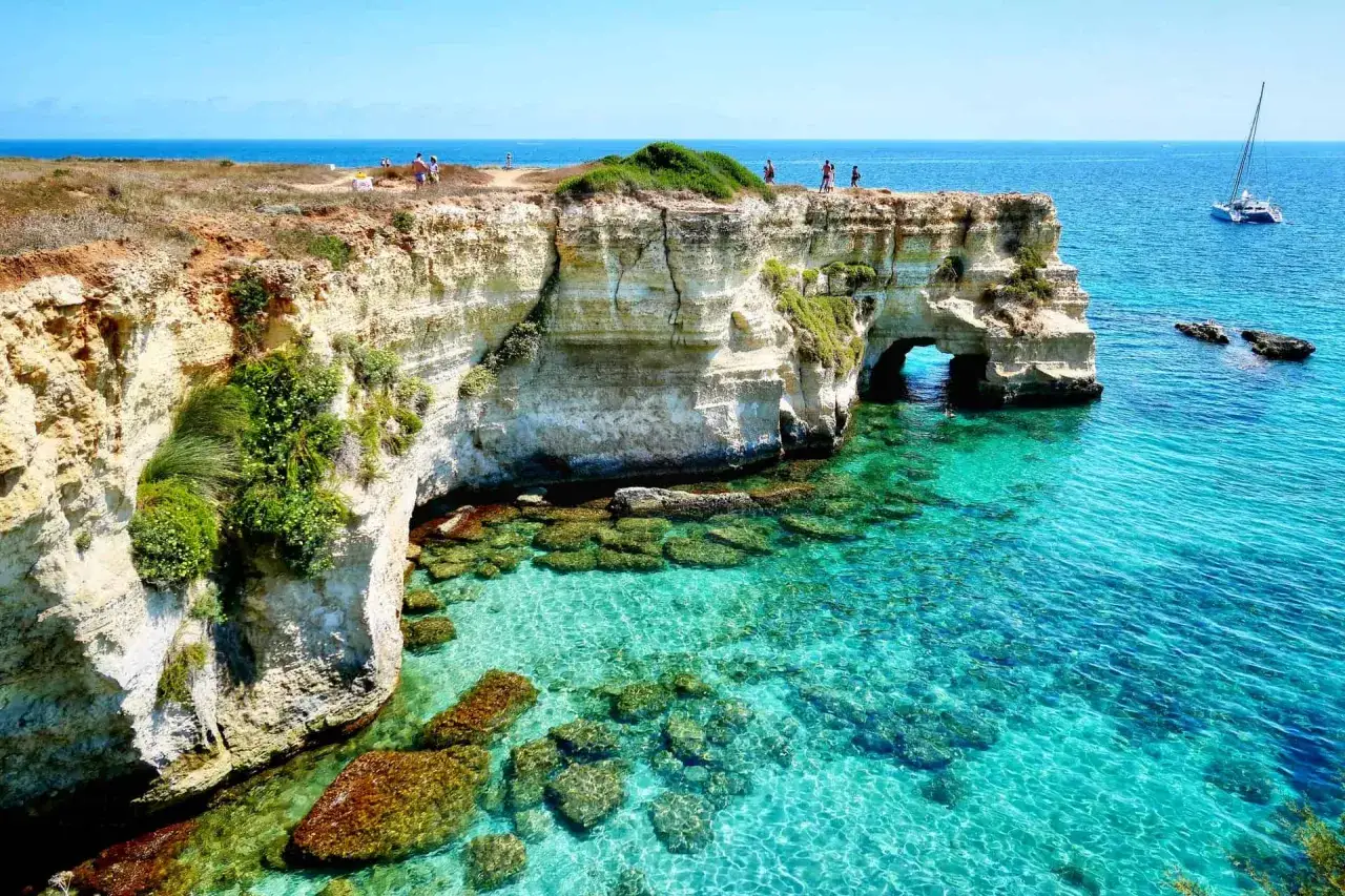 Scogliere bianche e acque cristalline sul litorale adriatico. Un arco naturale scolpito dal mare, con persone che ammirano il panorama e un catamarano all'orizzonte.