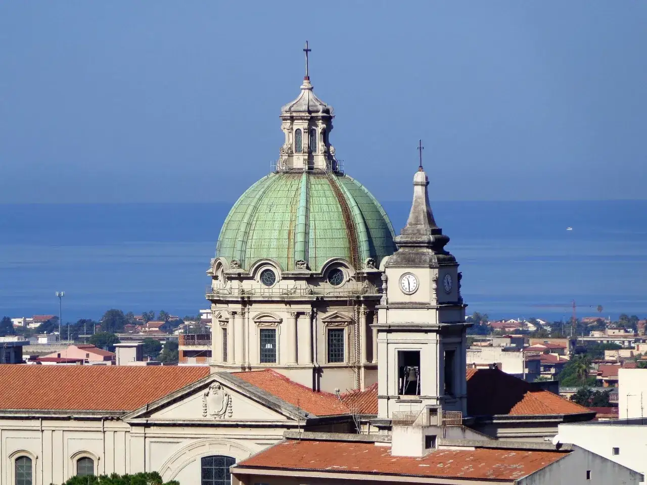 Cupola verde e campanile con orologio a Barcellona Pozzo di Gotto, con il mare azzurro sullo sfondo.