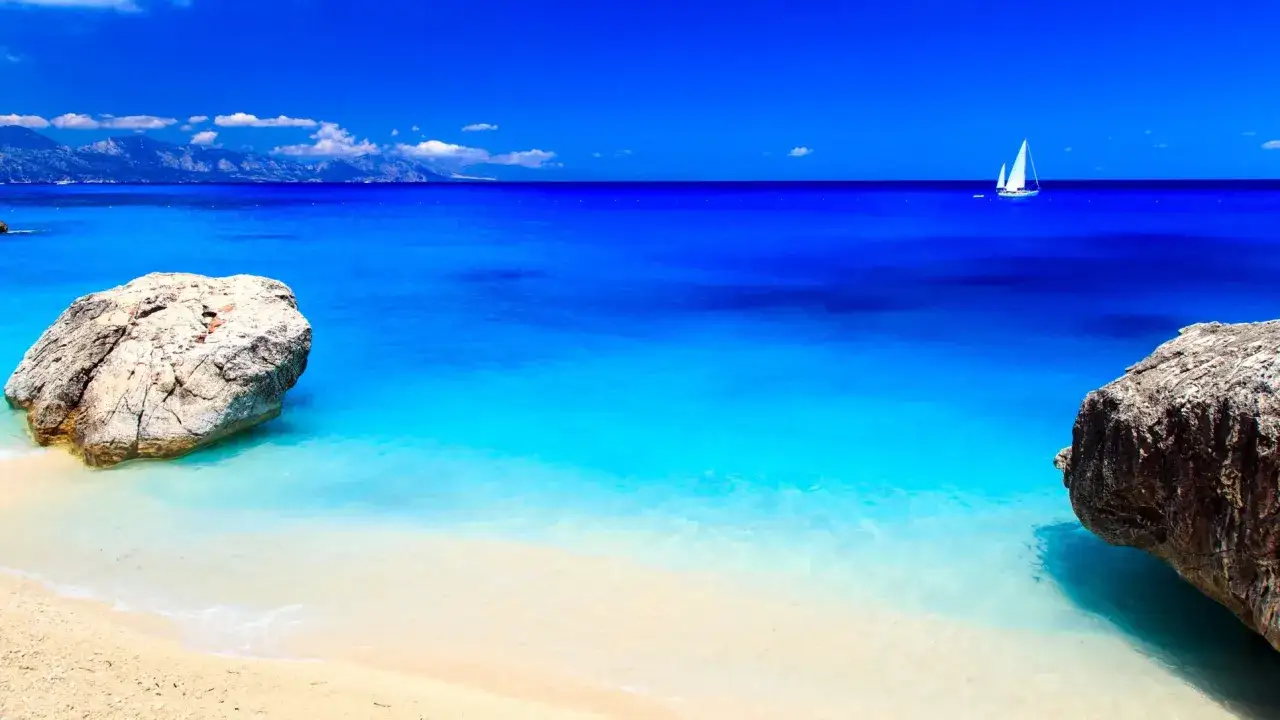 Spiaggia con rocce e un veliero in un mare bello in Italia. Acque cristalline e cielo azzurro intenso.