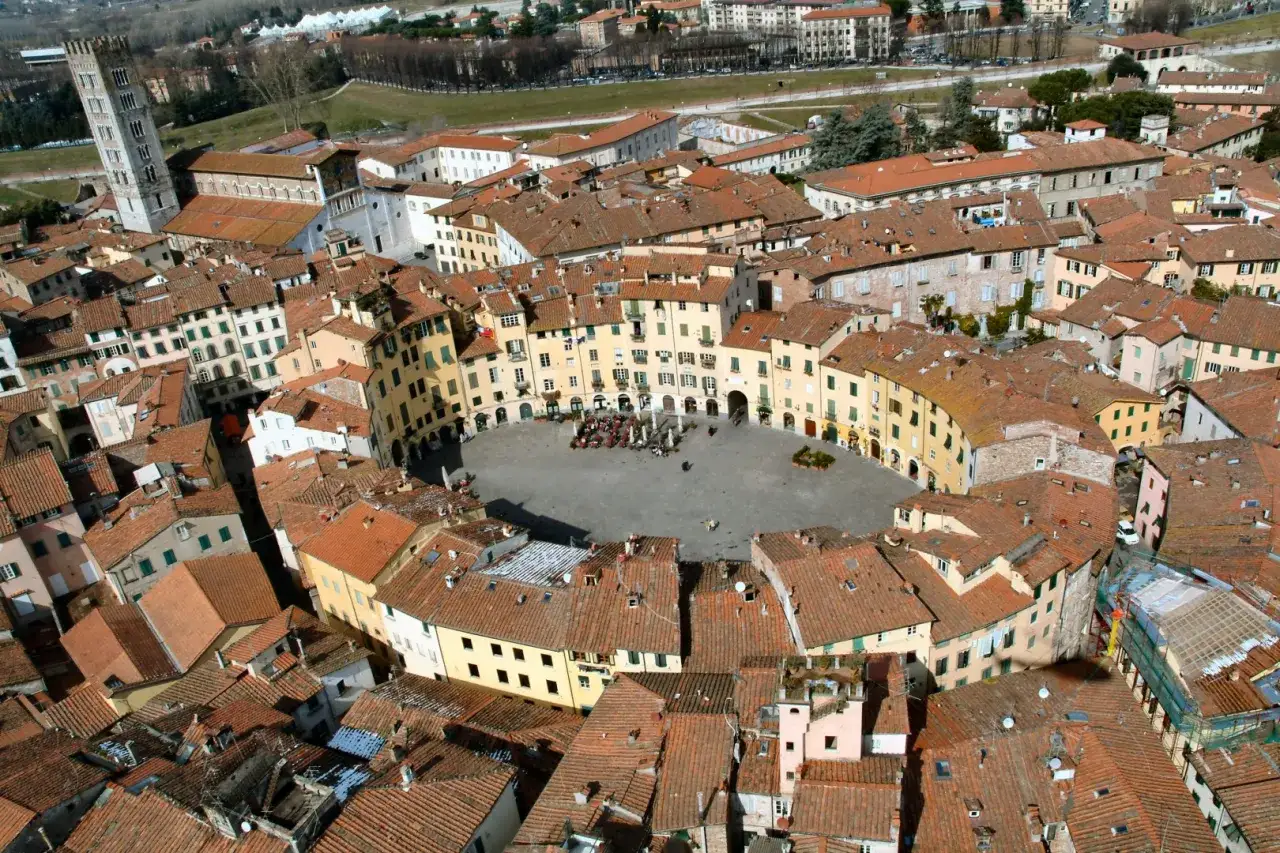 Piazza dell'Anfiteatro a Lucca, un gioiello da scoprire. Edifici ovali color pastello circondano la piazza, con la torre Guinigi in lontananza.