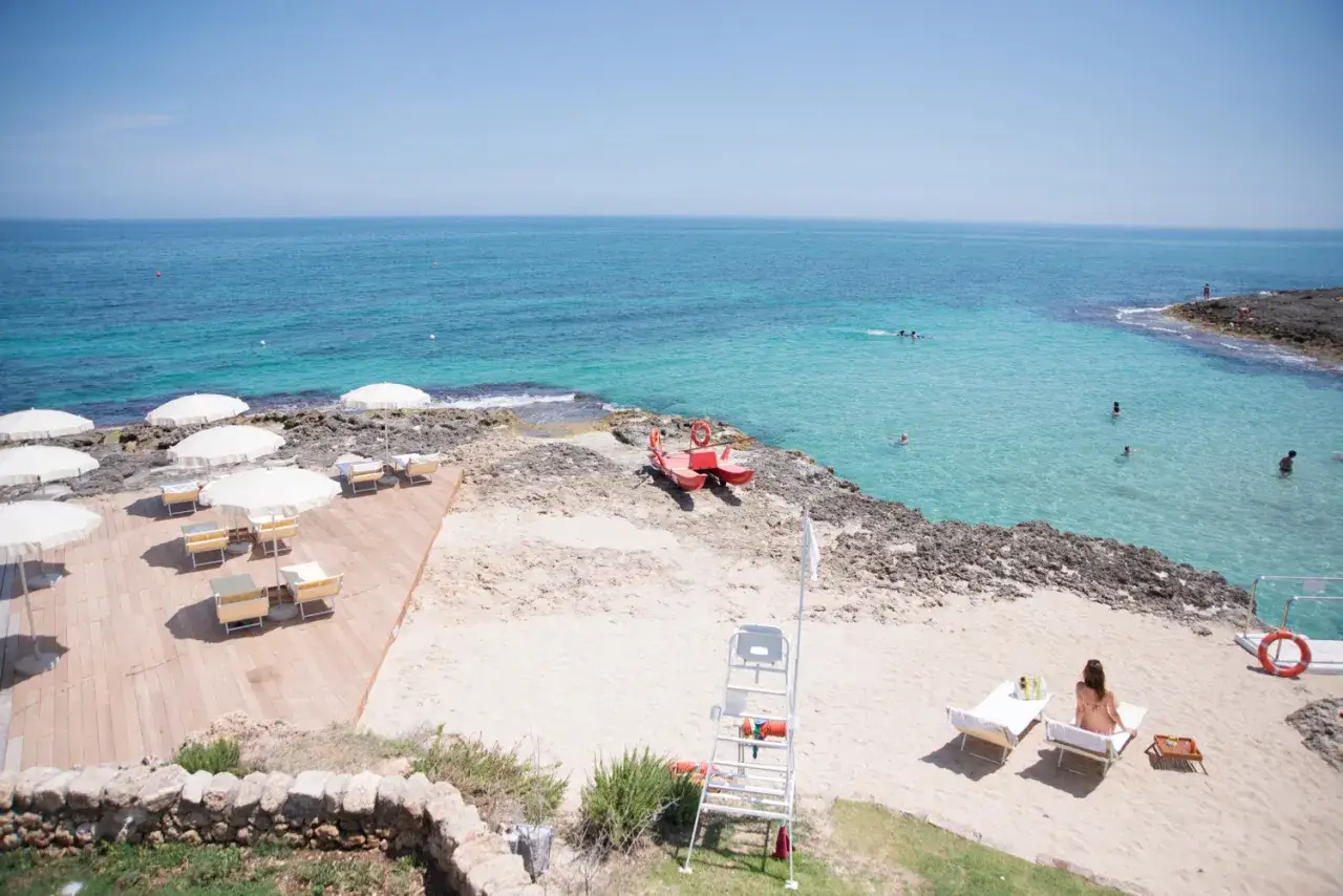 Lido Cala Pescatore: lettini, ombrelloni bianchi su pedana in legno e sabbia, mare cristallino con bagnanti, barca rossa di salvataggio.