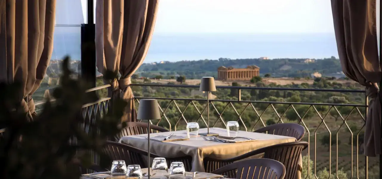 Tavoli apparecchiati su la terrazza sulla valle, con vista su un antico tempio greco e il mare all'orizzonte.