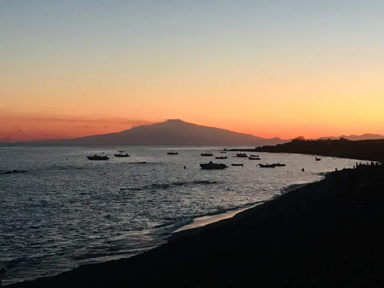 Tramonto sulla costa ionica calabrese, con barche all'ancora e una sagoma di montagna all'orizzonte.
