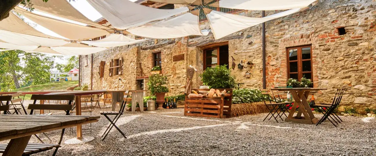 Cortile di agriturismo Cascina Mirandola con tavoli e sedie sotto un pergolato di teli bianchi. Le recensioni lodano l'atmosfera rustica.