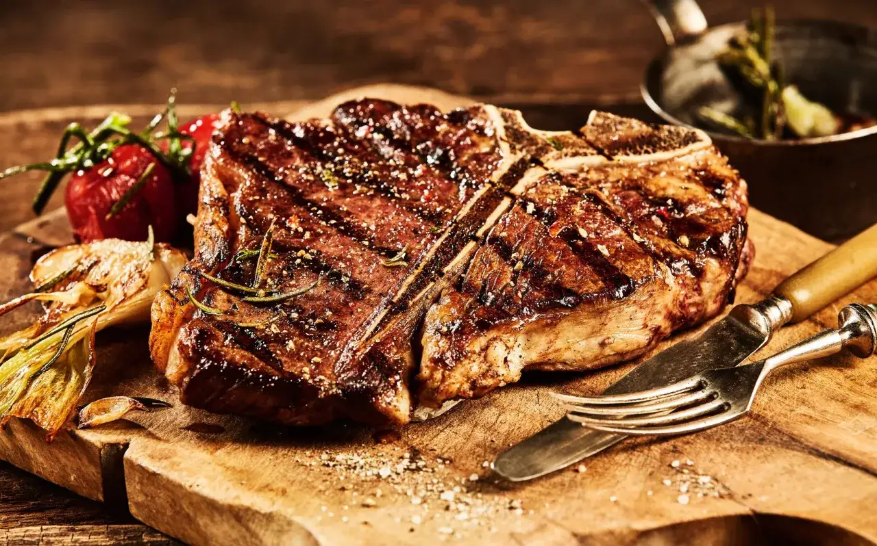 Una succulenta fiorentina, cotta alla perfezione, riposa su un tagliere di legno, accompagnata da pomodorini e aglio arrostiti.