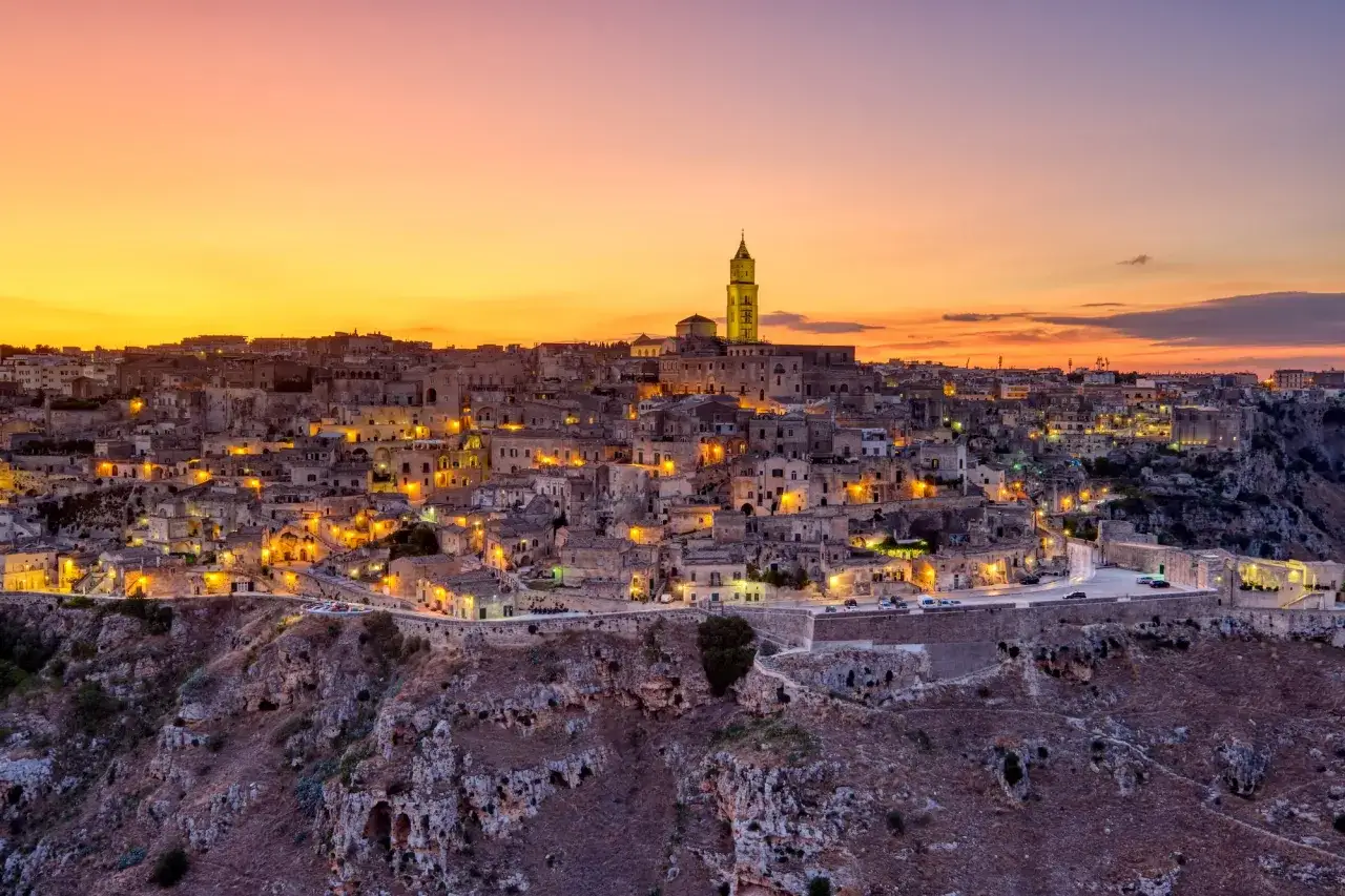 Matera illuminata al tramonto, un sogno da visitare in un giorno. Case antiche e un campanile svettano tra le rocce.