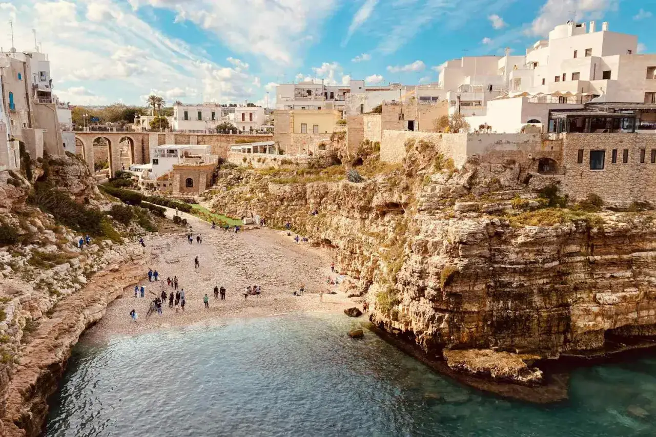 Scogliera a picco sul mare cristallino, con case bianche arroccate e una spiaggia di ciottoli animata da bagnanti. Un angolo di Puglia mare da sogno.