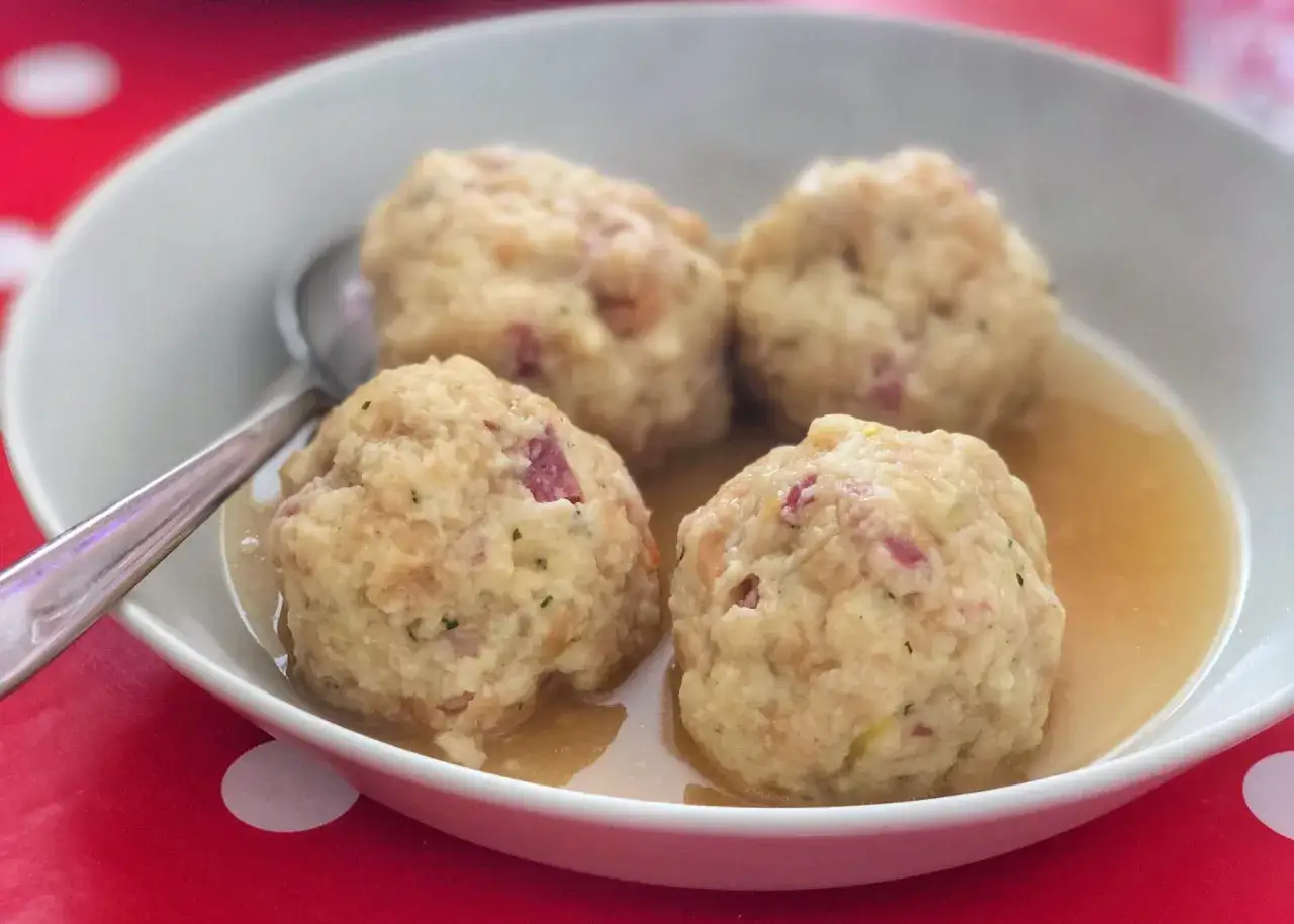 Canederli in brodo, un classico dei piatti tipici trentino alto adige, serviti in una ciotola bianca su un tavolo rosso a pois bianchi.
