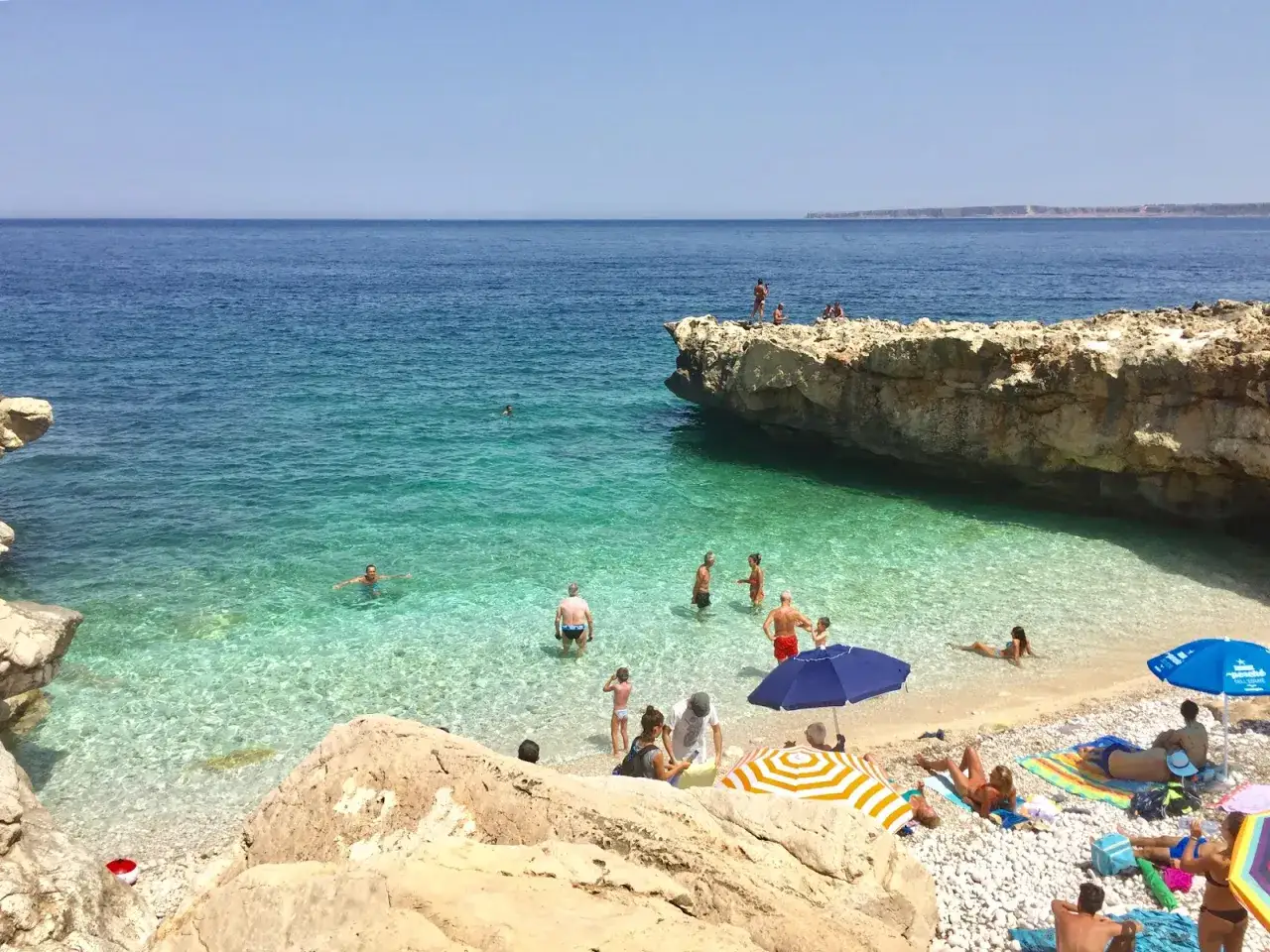 Persone si godono il sole e il mare cristallino sulla spiaggia di Guidaloca, tra scogli e acque turchesi.