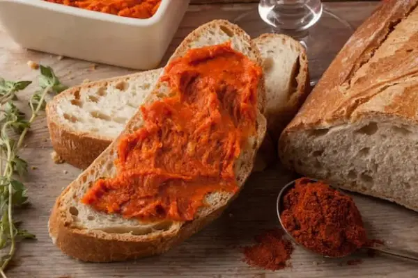 Fetta di pane con 'nduja, uno dei piatti tipici della Calabria, accanto a una ciotola con la crema e un cucchiaino di peperoncino in polvere.