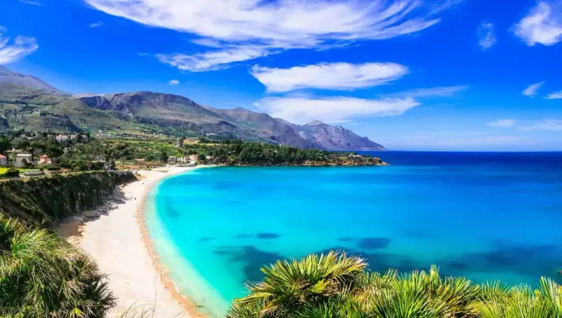 Una baia incantevole con acque cristalline e sabbia dorata, tra le spiagge più belle in Sicilia. Montagne verdi sullo sfondo.